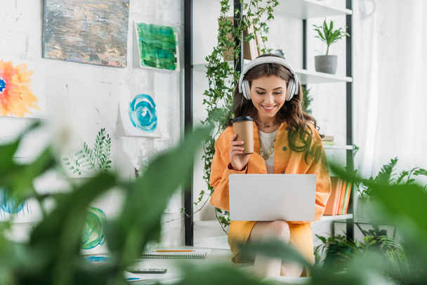 вибірковий фокус щасливої молодої жінки з паперовою чашкою прослуховування музики в навушниках і використання ноутбука
 - Фото, зображення