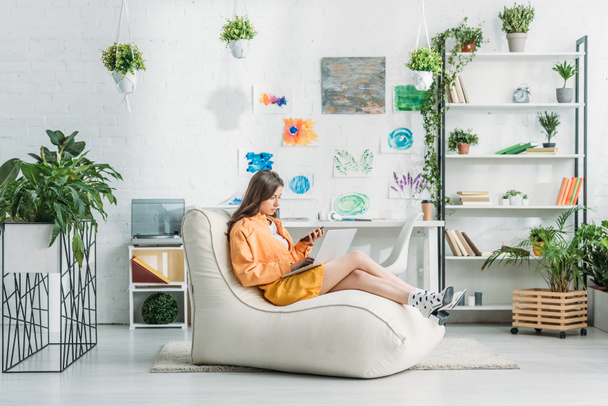 giovane donna che utilizza laptop e smartphone mentre è seduta su una morbida chaise lounge in camera spaziosa
  - Foto, immagini