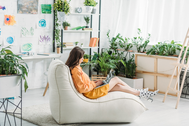 young woman using laptop while sitting on soft chaise lounge in light spacious room  - 写真・画像