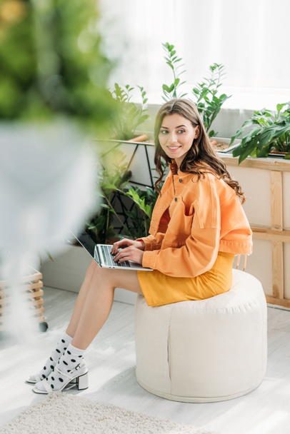 селективное внимание счастливой молодой женщины, сидящей на пуфе и использующей ноутбук дома
 - Фото, изображение