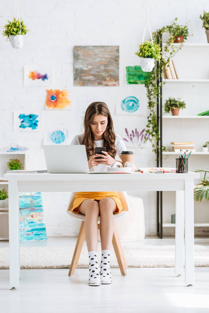 jonge vrouw met behulp van smartphone terwijl zitten aan het bureau in ruime kamer met schilderijen aan de muur - Foto, afbeelding