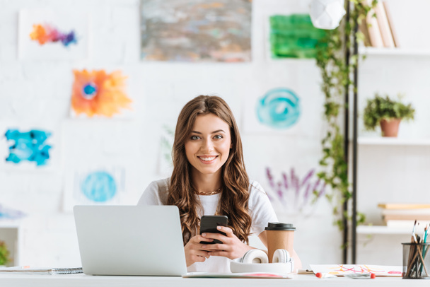 cheerful woman looking at camera while sitting at desk near laptop and using smartphone - Photo, Image