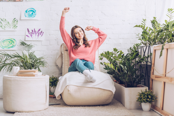 lachende meisje stretching terwijl zittend op zachte Chaise Lounge in de buurt van poef en weelderige groene planten - Foto, afbeelding