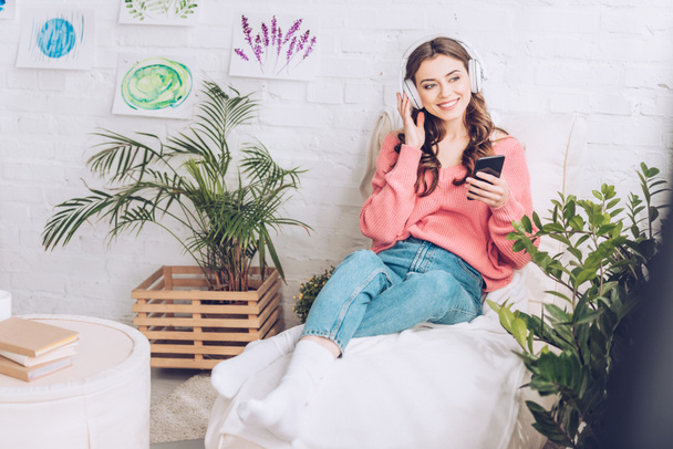 neşeli kız kulaklık müzik dinleme ve yumuşak şezlong dinlenme sırasında akıllı telefon kullanarak - Fotoğraf, Görsel
