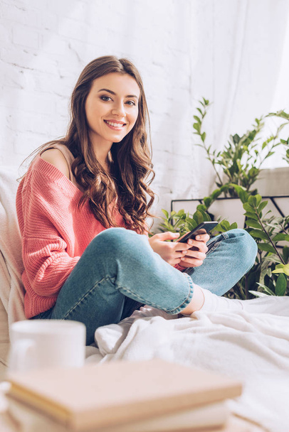selective focus of beautiful girl smiling at camera and using smartphone while sitting with crossed legs at home - Photo, Image