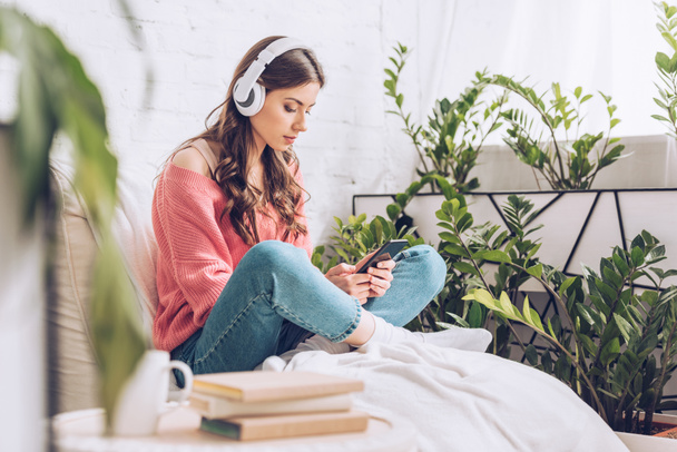 selective focus of pensive young woman sitting with crossed legs, listening music in headphones and using smartphone - Foto, immagini