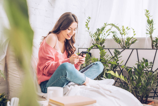 selective focus of cheerful girl using smartphone while sitting with crossed legs near green plants at home - Photo, Image