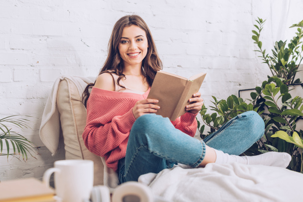 привлекательная веселая девушка улыбается в камеру, сидя с книгой на мягком шезлонге
 - Фото, изображение