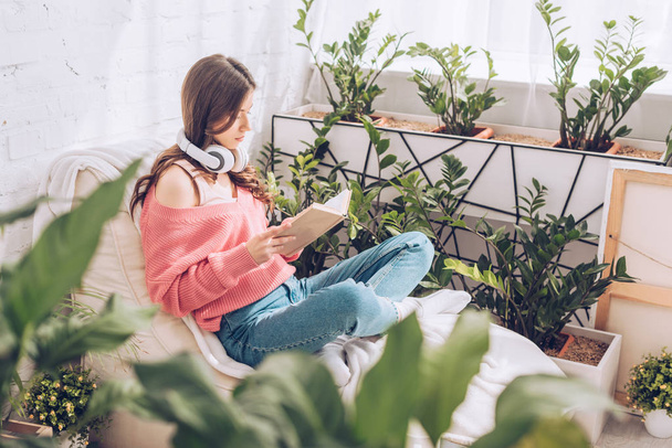 selective focus of young woman reading book while sitting with crossed legs surrounded by lush plants at home - Zdjęcie, obraz