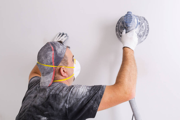 Plasterer wearing dust mask polishes a wall with sanding machine . House renovation concept. - Foto, immagini