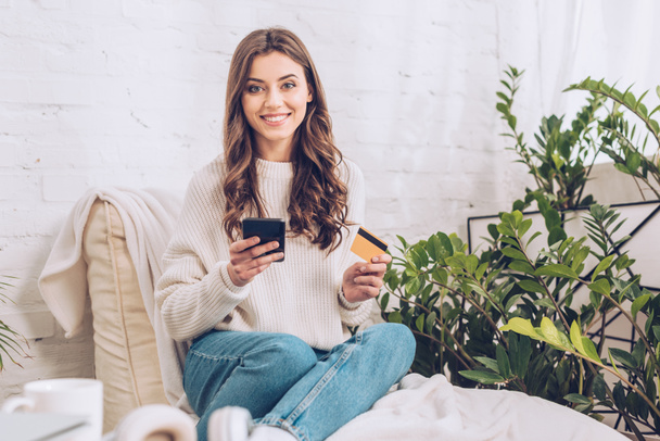 happy pretty girl holding credit card and using smartphone while smiling at camera - Foto, Bild