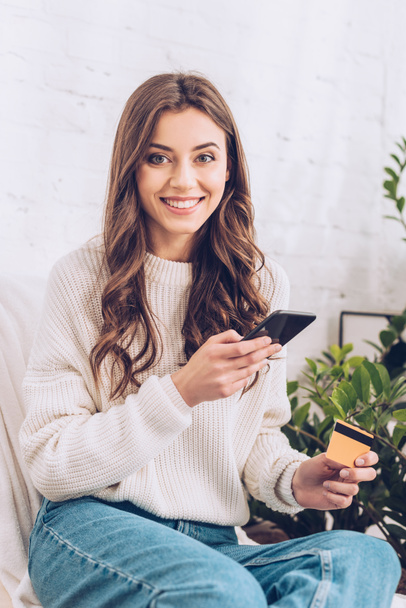happy young woman looking at camera while holding credit card and smartphone - Foto, afbeelding