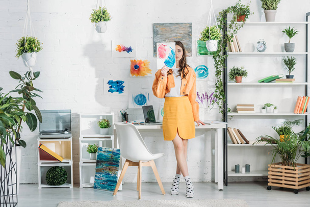 trendy young woman covering face with painting while standing in spacious room decorated with green plants and drawings on white wall - Foto, afbeelding