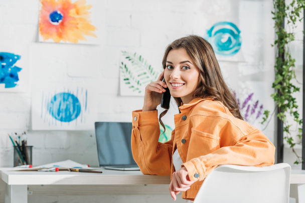 vidám lány mosolyogva a kamera mellett ülve íróasztal fal színes festmények - Fotó, kép