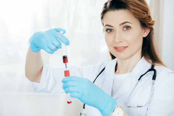血液サンプルと試験管で注射器を保持するラテックス手袋で笑顔の医師  - 写真・画像