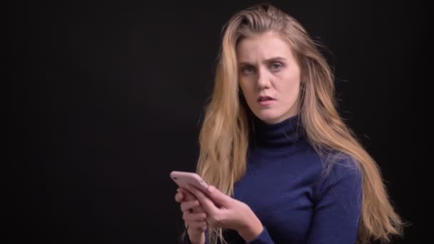 Young blonde model working with smartphone raises glance to camera and smiles into it on black background. - Video