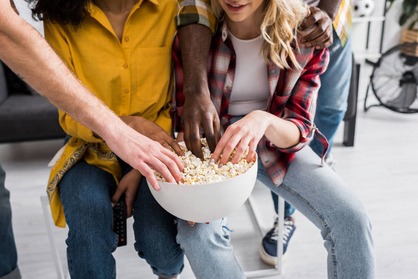 Vista ritagliata di quattro amici multiculturali con ciotola di popcorn in soggiorno
 - Foto, immagini