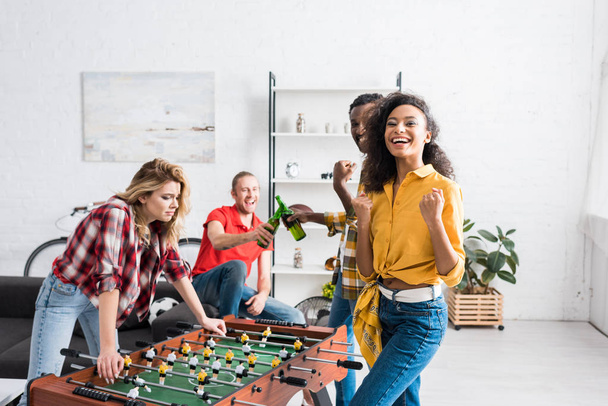 Iloiset ja hymyilevät monikansalliset miehet ja naiset pelaavat pöytäjalkapalloa olohuoneessa kotona
 - Valokuva, kuva