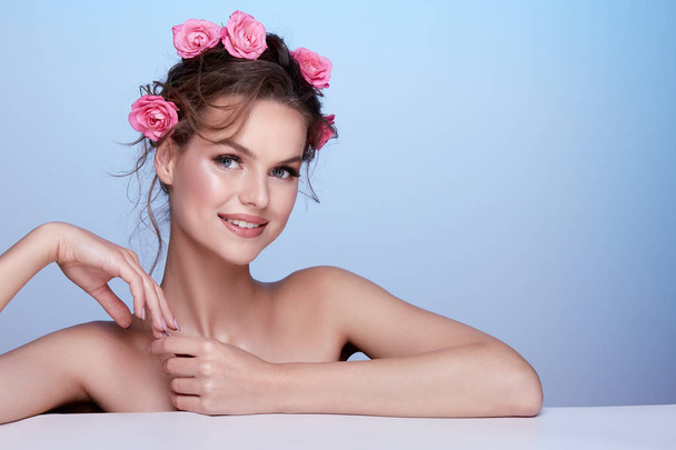 πορτραίτο της ελκυστικής νεαρής γυναίκας με ροζ τριαντάφυλλα στα μαλλιά που ποζάρουν σε μπλε φόντο - Φωτογραφία, εικόνα
