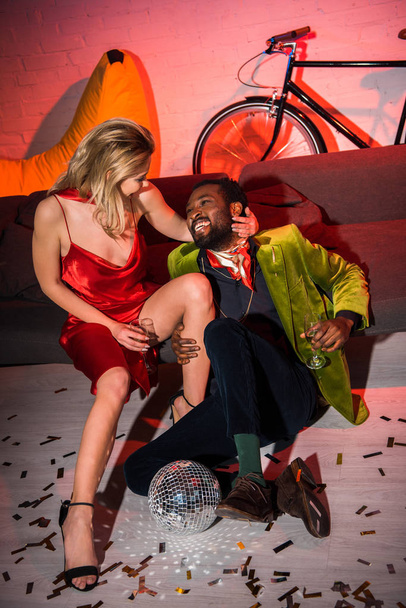 joven rubia en vestido rojo sentado con hombre afroamericano en club nocturno
  - Foto, imagen
