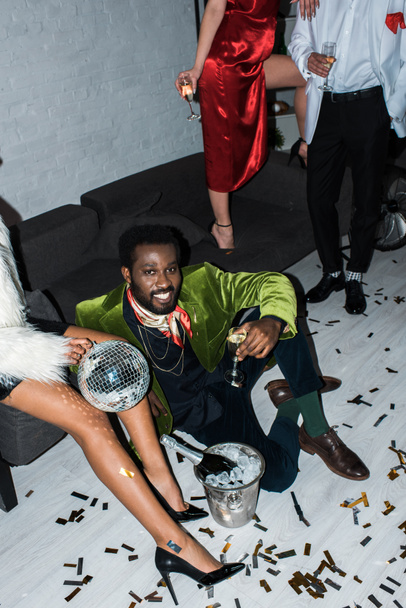 Widok napowietrznych African American mężczyzna trzyma kieliszek do szampana, siedząc na podłodze w pobliżu przyjaciół  - Zdjęcie, obraz
