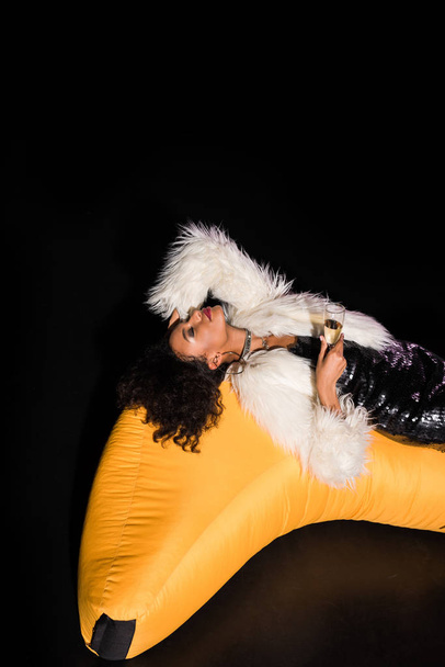 gai afro-américaine fille couché sur chaise sac haricot jaune et tenant verre de champagne sur noir
  - Photo, image
