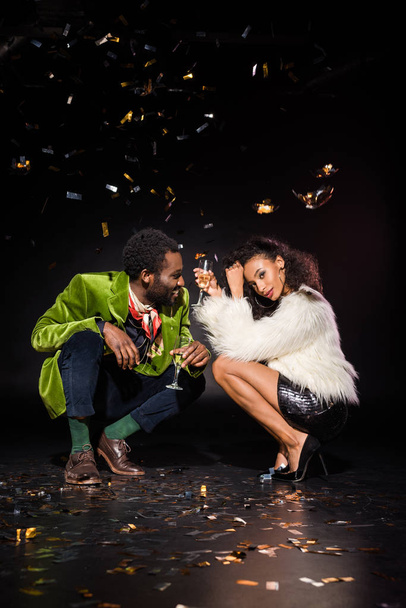 vrolijke Afro-Amerikaanse man en mooie Afro-Amerikaanse vrouw die Champagne glazen vasthoudt terwijl ze in de buurt van vallende confetti op zwart zitten  - Foto, afbeelding