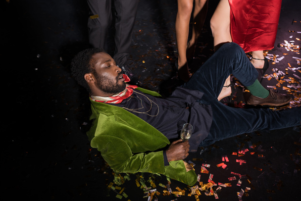ハンサムで酔っぱらったアフリカ系アメリカ人男性が黒の友人の近くに紙吹雪と床に横たわっている  - 写真・画像