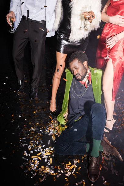 handsome and drunk african american man lying on floor with shiny confetti near friends on black  - Photo, Image