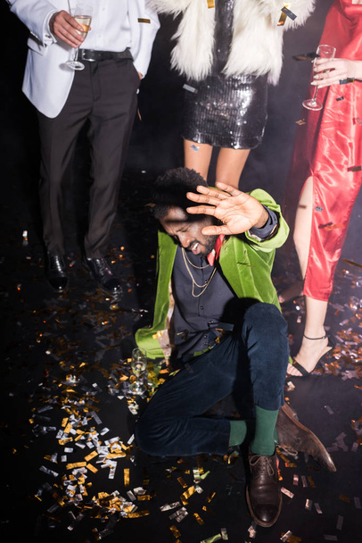 drunk african american man with closed eyes gesturing while lying on floor with shiny confetti near friends on black  - Foto, afbeelding