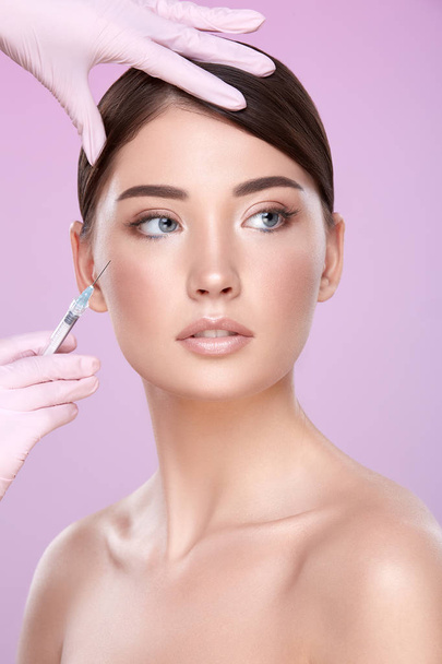 partial view of cosmetologist in rubber gloves with syringe and young brunette woman on pink background - Photo, image