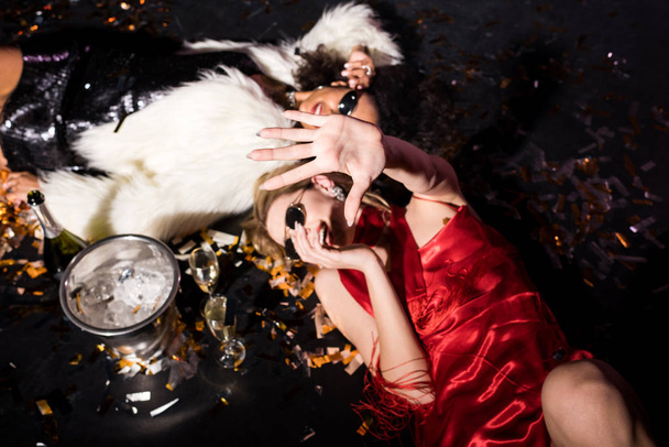 overhead view of blonde girl in red dress gesturing while smiling and lying on floor with confetti near african american friend on black  - 写真・画像