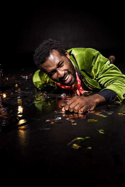 selective focus of emotional and drunk african american man lying on floor with shiny confetti on black  - Photo, Image