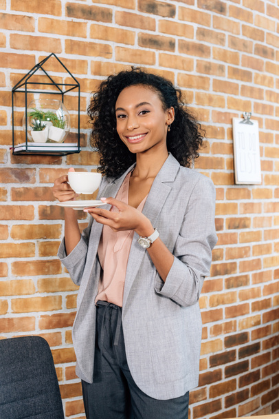 belle souriante afro-américaine femme d'affaires occasionnelle tenant tasse de café avec soucoupe au bureau
 - Photo, image
