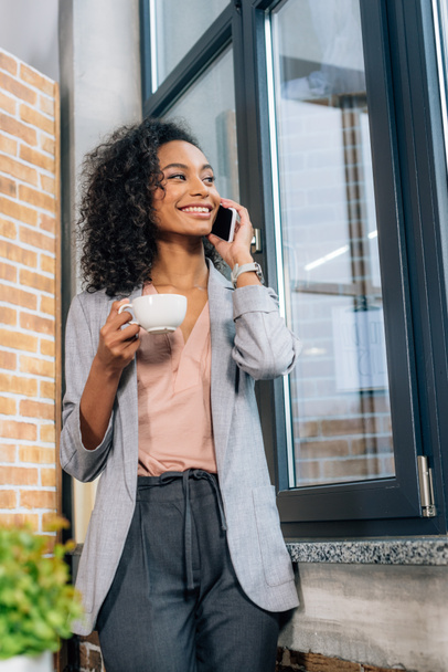 Afrikanisch-amerikanische Gelegenheitsunternehmerin mit Kaffeetasse spricht im Loft-Büro auf dem Smartphone - Foto, Bild