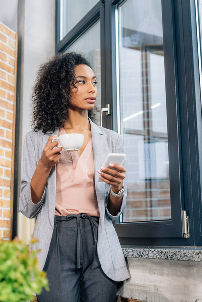 african american Casual businesswoman with coffee cup using smartphone in loft office - Zdjęcie, obraz