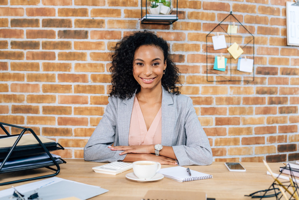 hermosa sonriente ocasional afroamericana mujer de negocios sentada en el escritorio en la oficina loft
 - Foto, Imagen