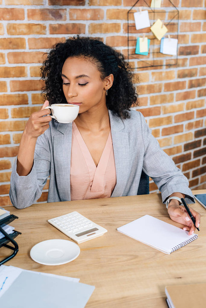 Afrikanisch-amerikanische Gelegenheitsunternehmerin am Schreibtisch beim Kaffeetrinken im Büro - Foto, Bild
