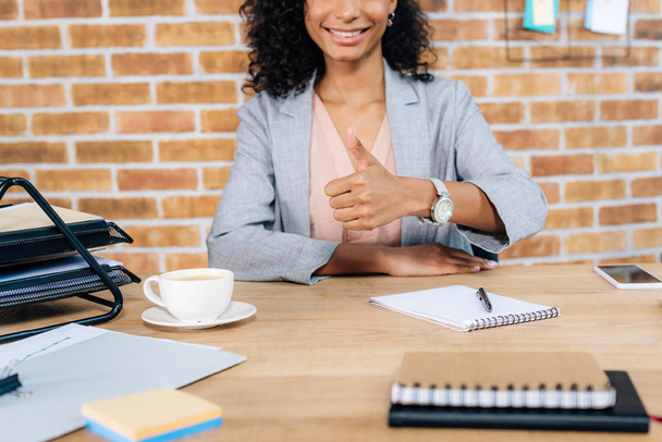 Przycięte widok uśmiechnięty casual African American businesswoman pokazując kciuka w górę w biurze - Zdjęcie, obraz