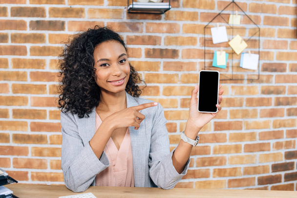 african american Casual businesswoman pointing with finger at smartphone with blank screen in loft office - Photo, Image