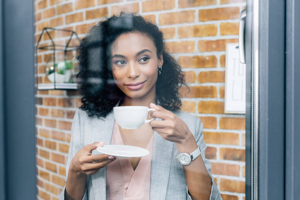 African American casual zakenvrouw met koffie kopje achter venster in Office - Foto, afbeelding