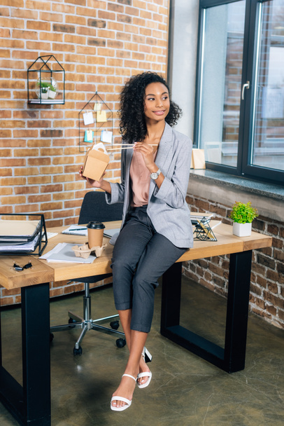 smiling african american Casual businesswoman sitting on desk with coffee to go and takeaway box  - Photo, Image