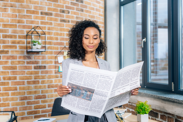 atractiva afroamericana Casual mujer de negocios leyendo periódico de negocios en la oficina loft
 - Foto, Imagen