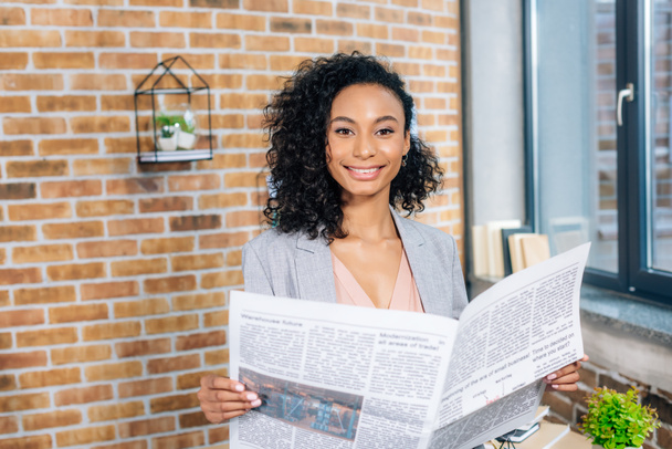krásná usmívající se africká americká příležitostná obchodžena s obchodními novinami v podkrovní kanceláři - Fotografie, Obrázek
