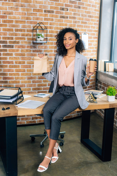 African American dorywczo businesswoman trzyma na wynos worek papierowy i kawę, aby przejść w biurze - Zdjęcie, obraz