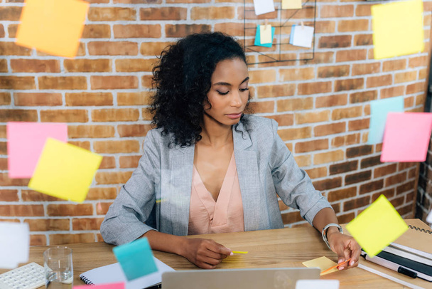 Casual african american businesswoman sitting at desk in front of glass window with Sticky Notes  - Фото, зображення