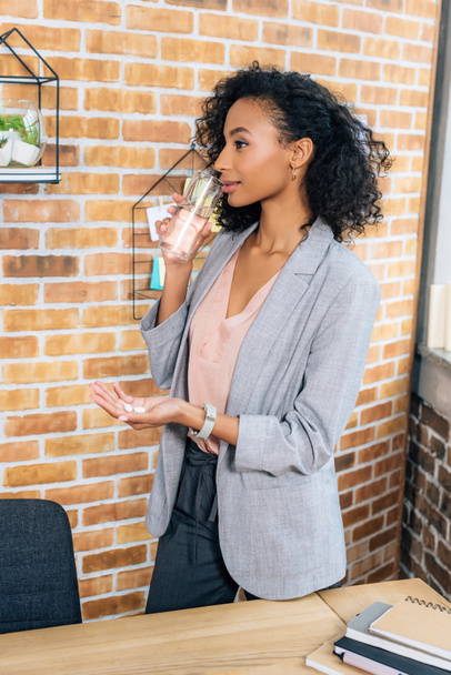 africano americano casual mujer de negocios con píldoras beber agua en oficina
  - Foto, imagen