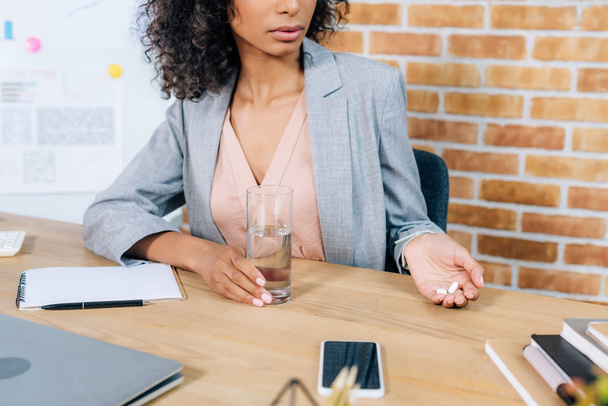 обрізаний вид афроамериканської випадкової бізнес-леді з таблетками і склянкою води за офісним столом
  - Фото, зображення