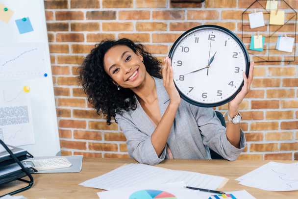 souriant afro-américain femme d'affaires occasionnelle tenant horloge dans le bureau loft
 - Photo, image