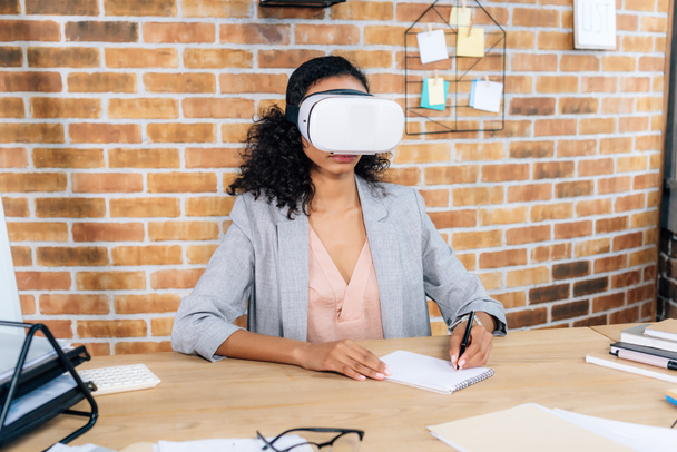 afroamericana Casual mujer de negocios en realidad virtual auriculares en escritorio de oficina escritura en cuaderno
 - Foto, Imagen